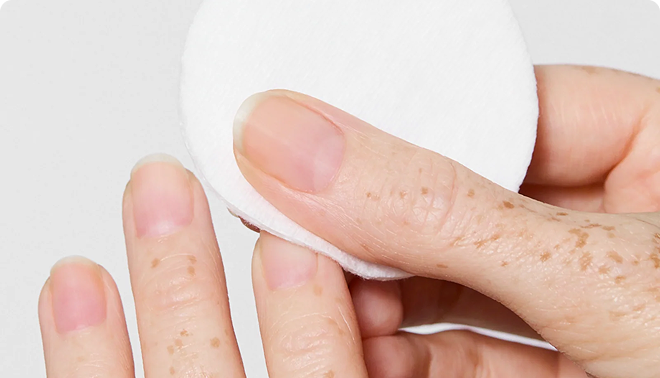 A close-up of a hand with freckles wiping fingernails with a white cotton pad against a white background.