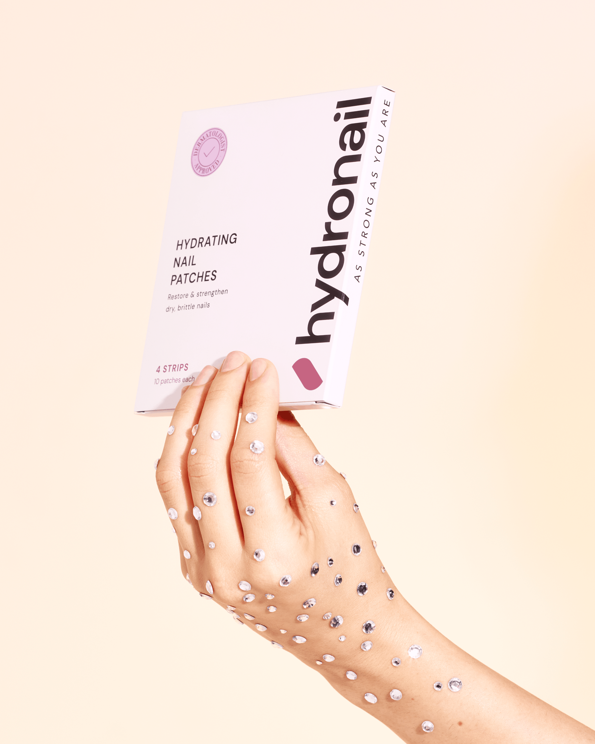 A hand adorned with rhinestones holds up a white box of Hydronail Hydrating Nail Patches.