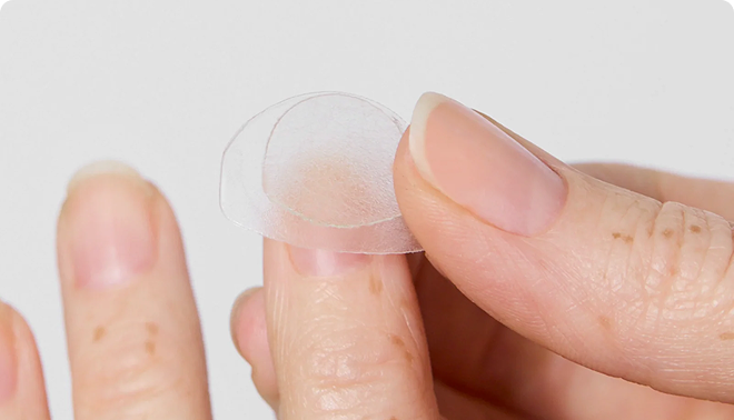 A close-up of a hand holding two small, round, translucent patches between the thumb and forefinger.