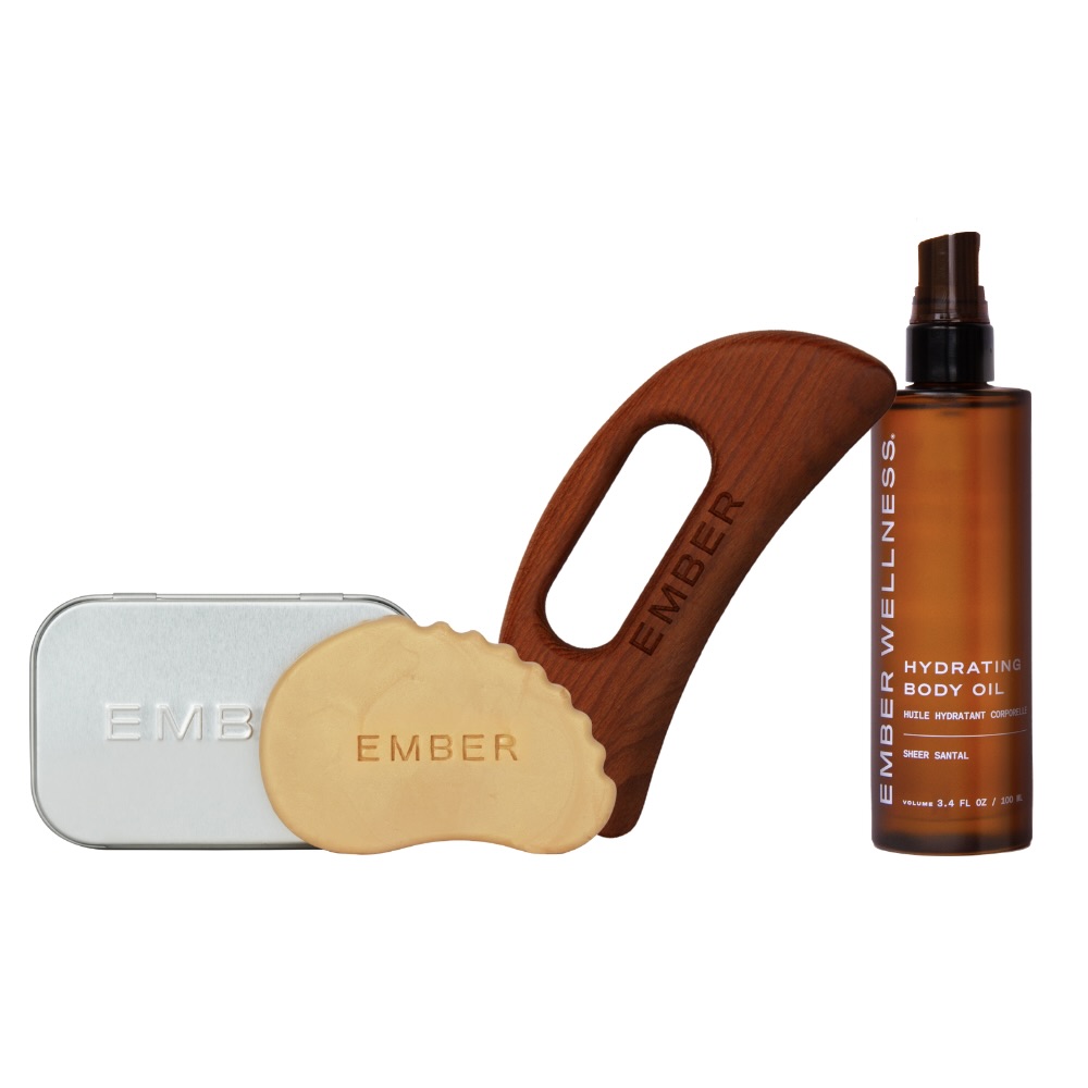 An Ember Wellness hydrating body oil bottle next to a wooden and stone gua sha tool with a tin.