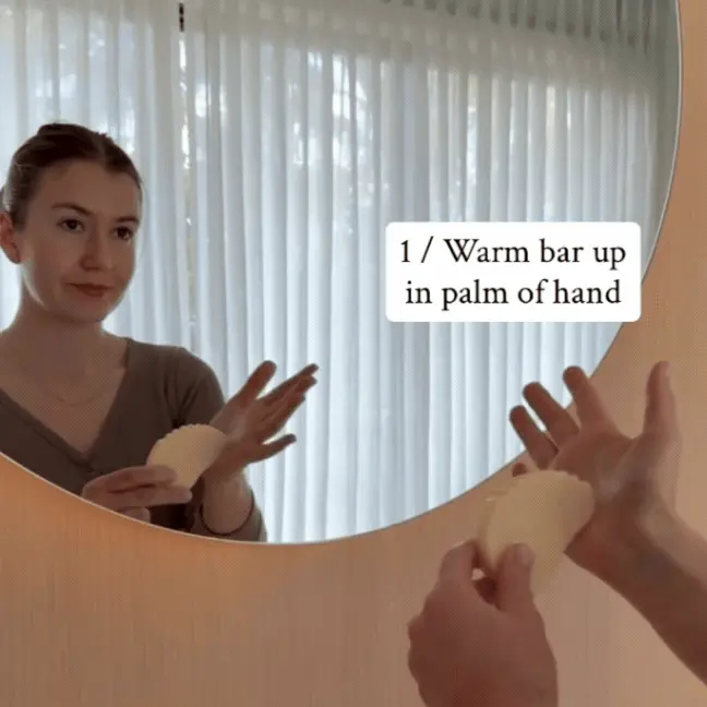A woman looks in a mirror, holding a scalloped bar with text overlay: 'Warm bar up in palm of hand.'