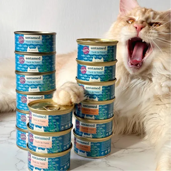 A large, fluffy cat yawns widely with its paw on two stacks of canned cat food.