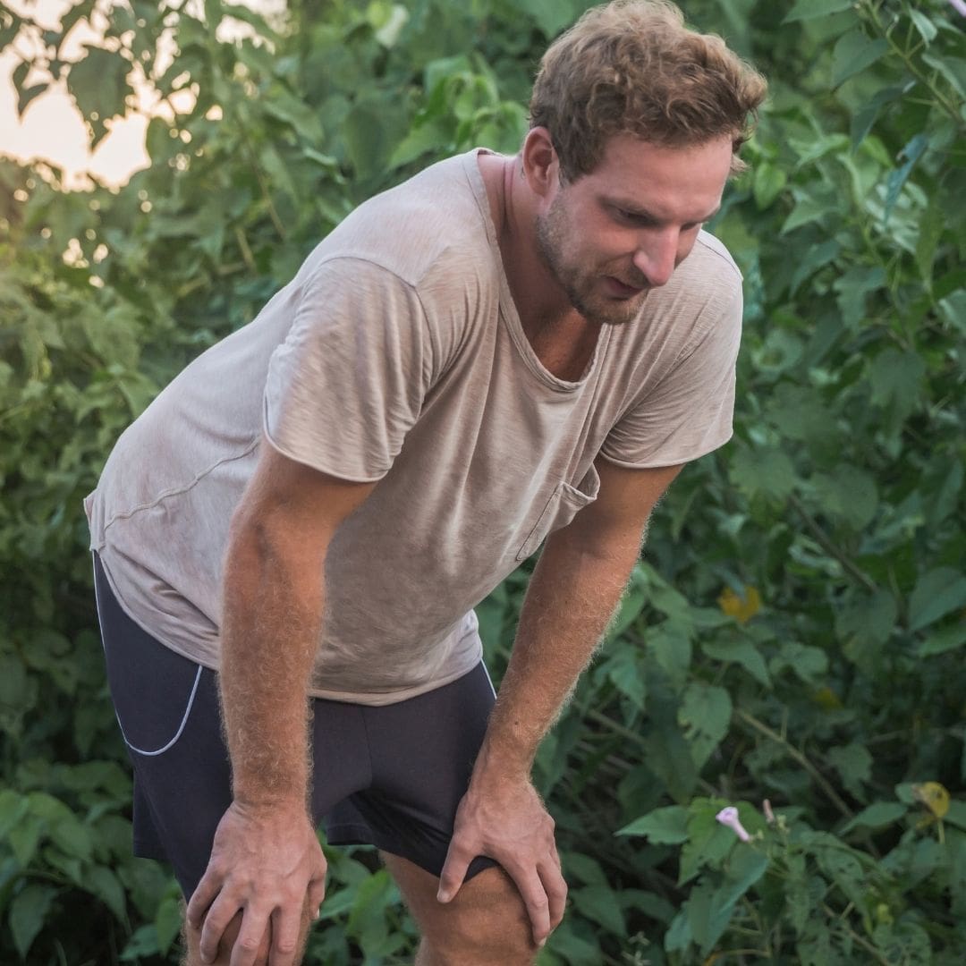 Man in a sweaty shirt bending over, surrounded by greenery.