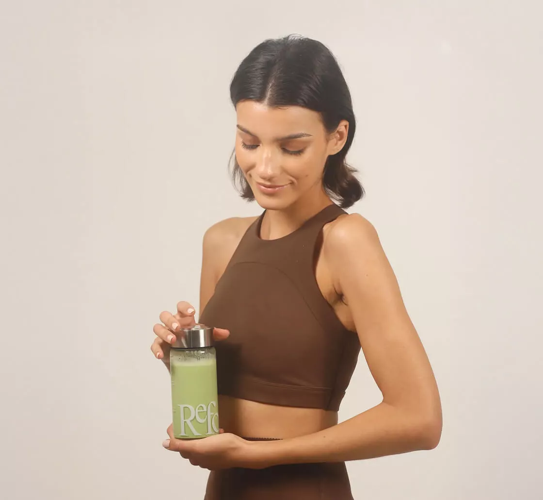 A woman in a brown workout top holds a glass bottle filled with a green smoothie.