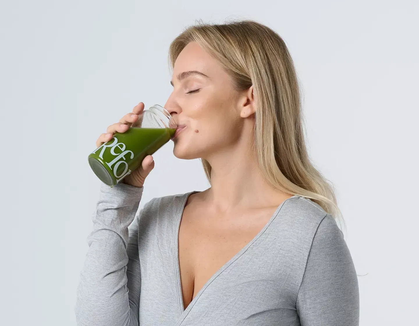 A blonde woman with her eyes closed drinks a green beverage from a glass bottle against a white background.