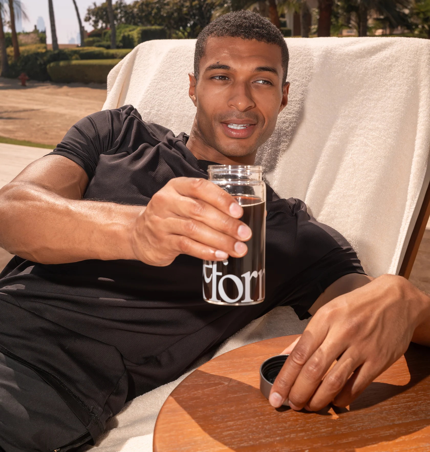 A man in a black t-shirt relaxes on a lounge chair outdoors holding a glass with a dark drink.
