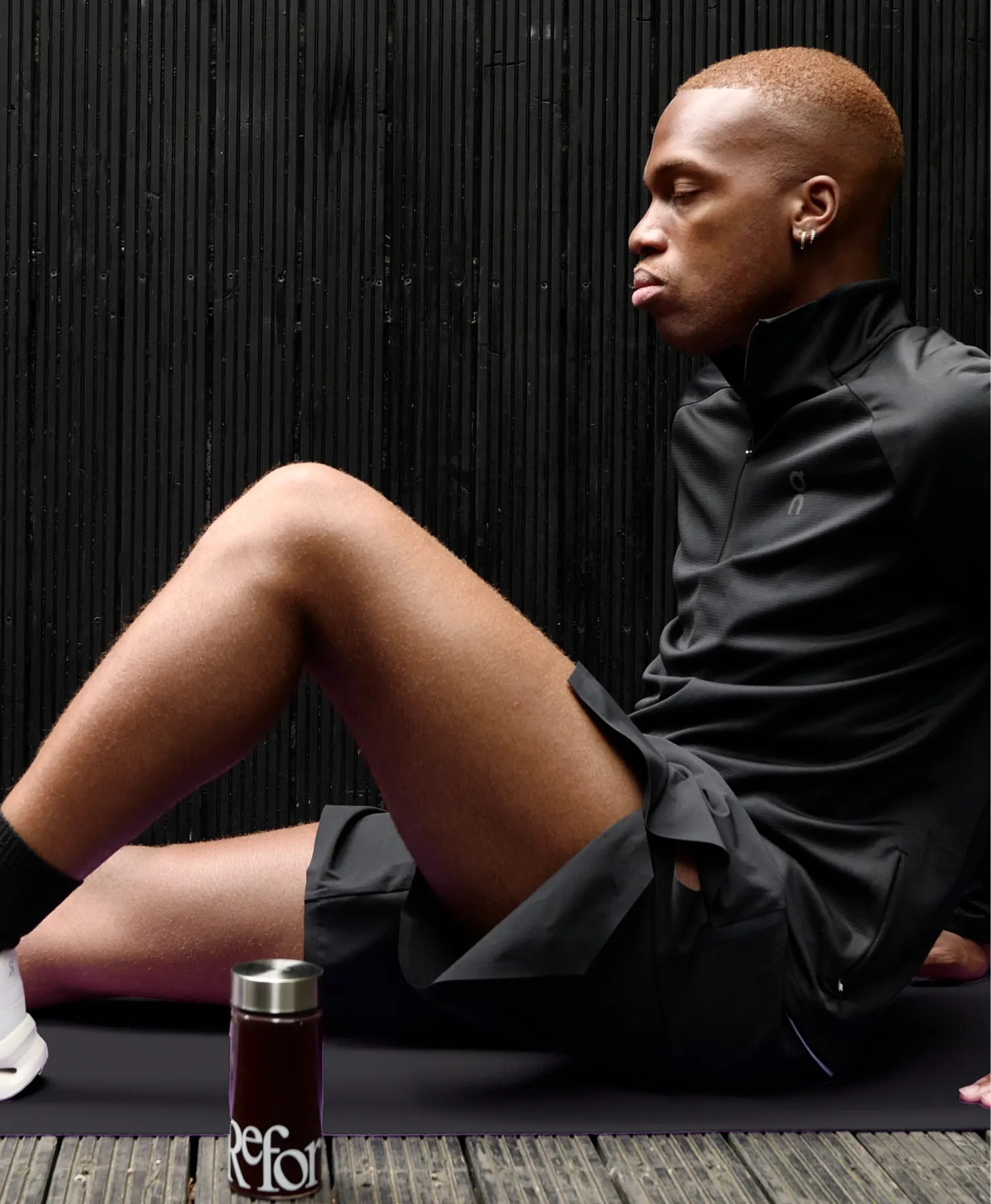 A man in black athletic clothing sits on a mat against a dark wall, resting with his eyes closed.