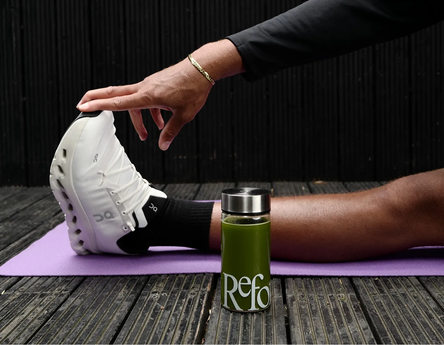 A person stretches on a purple yoga mat, reaching for their white sneaker next to a green drink.