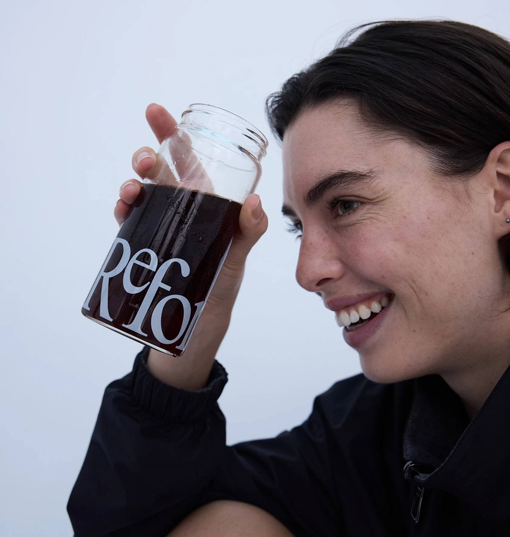 A close-up of a smiling woman in a black jacket holding a glass jar of a dark beverage.