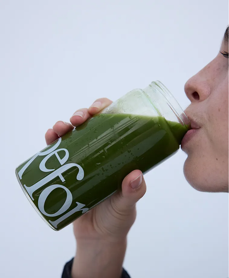 A close-up of a person drinking a green beverage from a glass tumbler with white text on it.