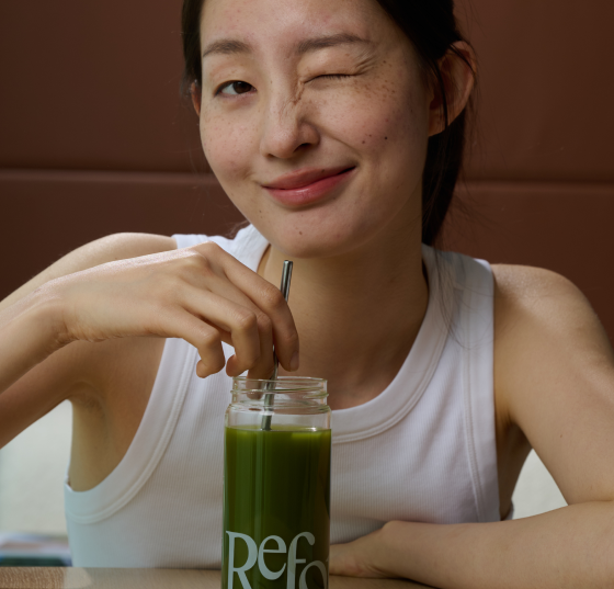 A woman in a white tank top winks at the camera while stirring a green drink.