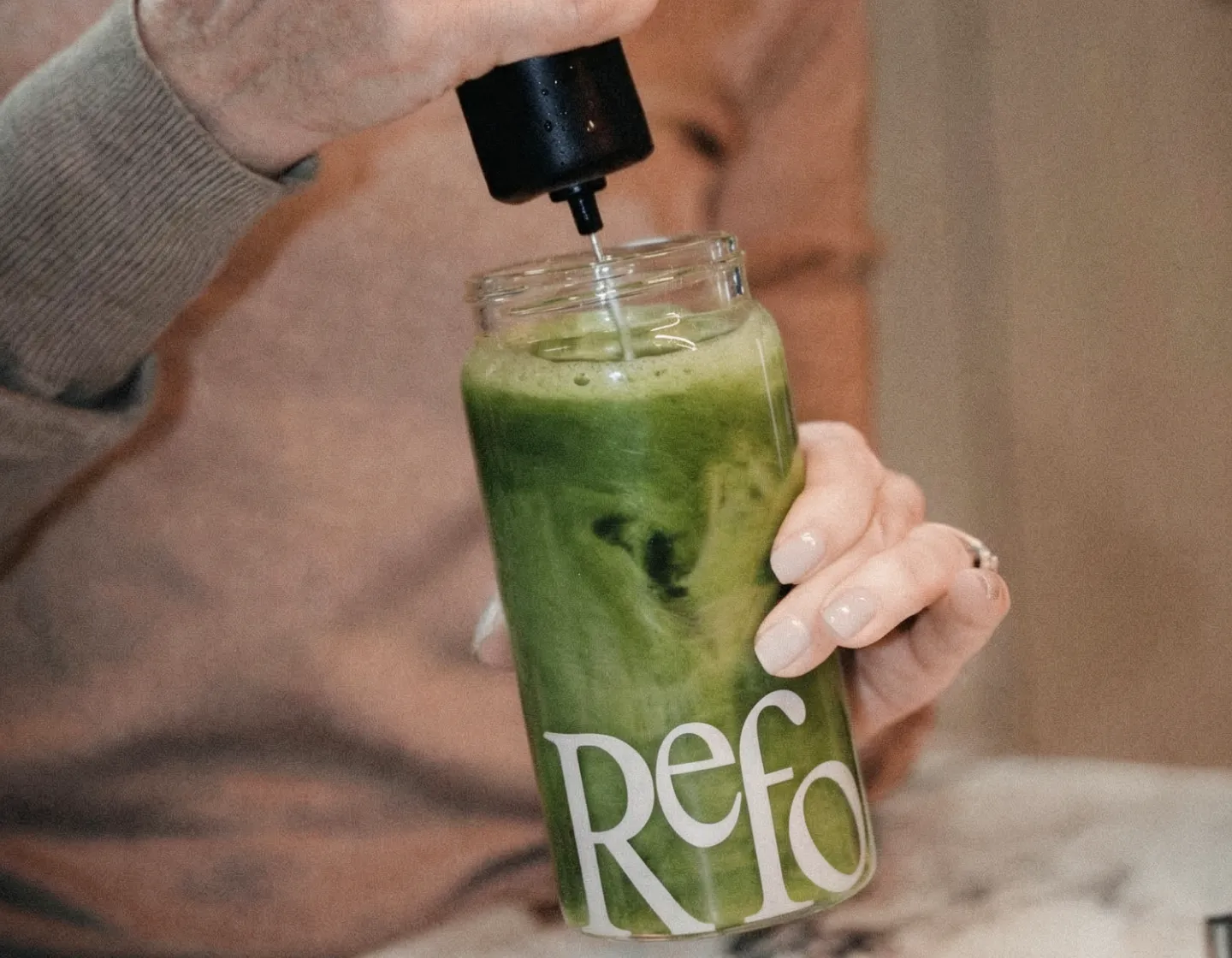 A person mixes a frothy green drink in a glass jar with a handheld frother.