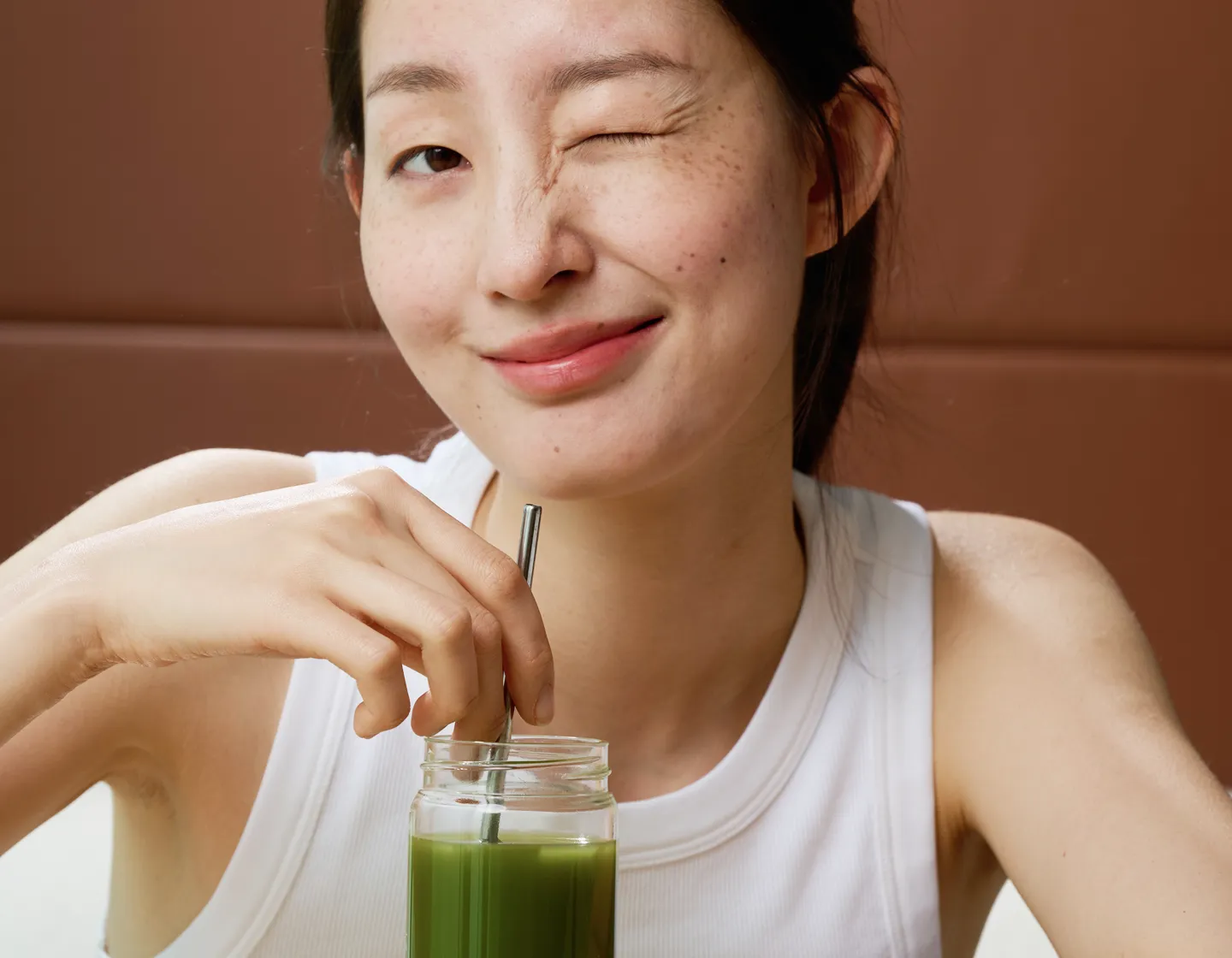 A smiling woman winks at the camera while stirring a green drink in a glass jar.