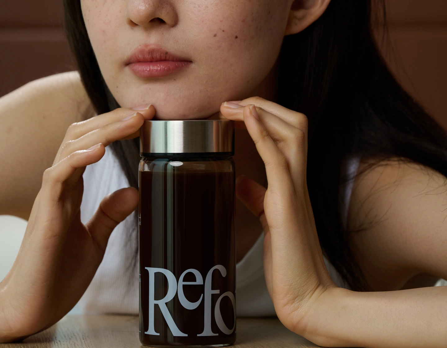 A woman rests her chin on her hands, holding a glass bottle filled with a dark liquid.