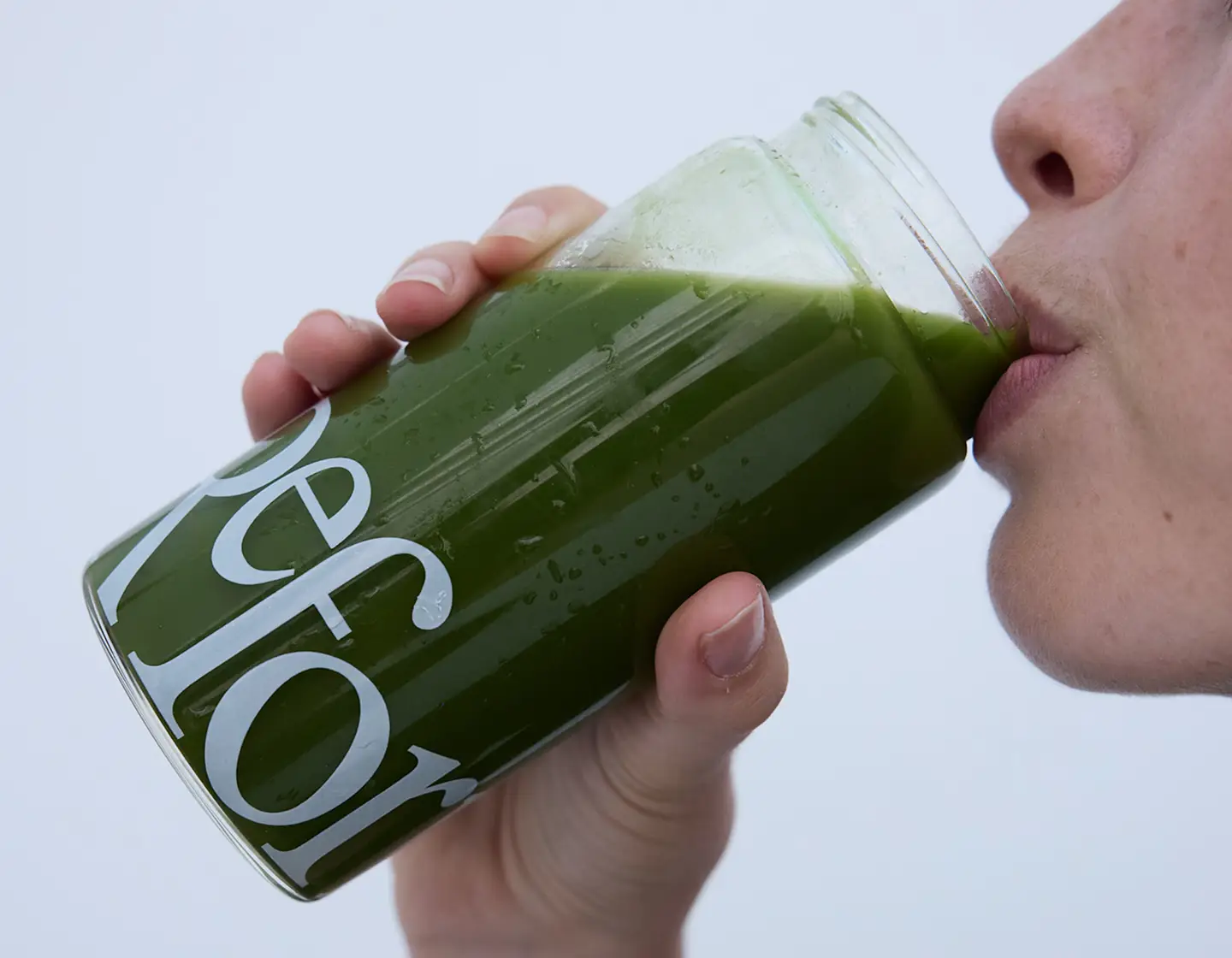 A close-up of a person drinking a green beverage from a glass jar with white lettering.