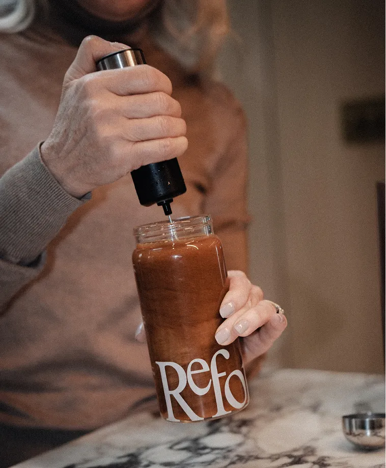 A person uses a handheld pump device to mix a brown beverage in a glass jar labeled 'Refo'.