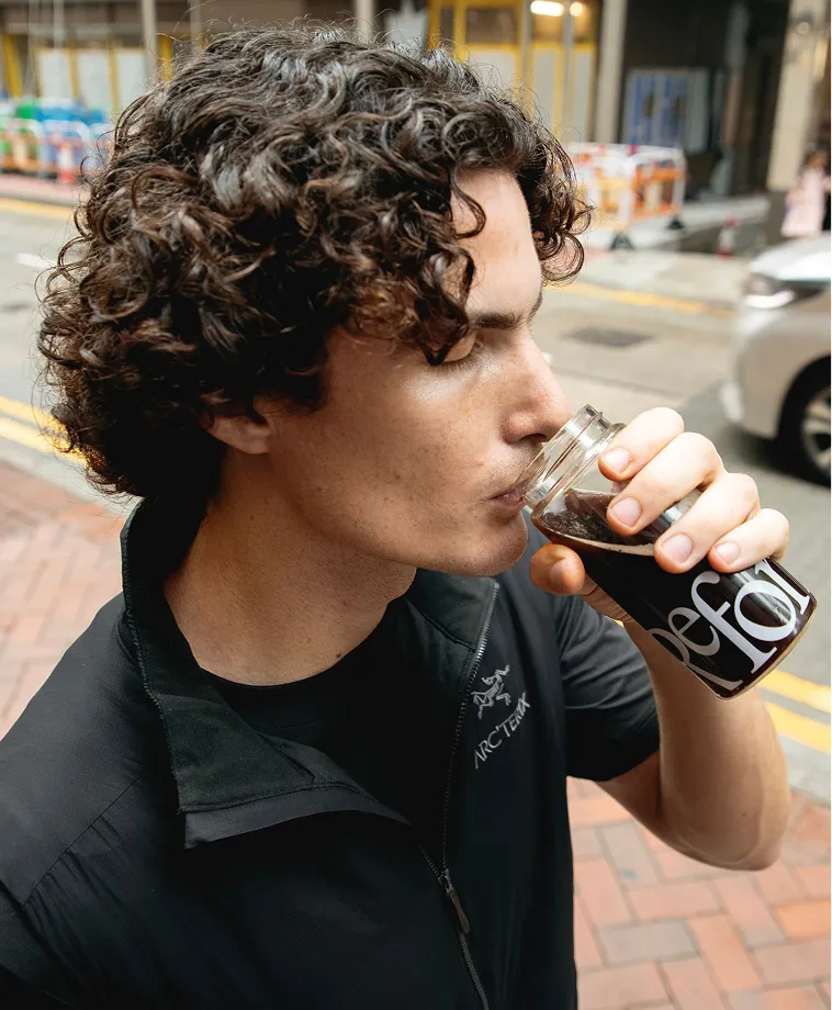 A man with curly hair wearing a black jacket sips a dark beverage from a glass jar outdoors.