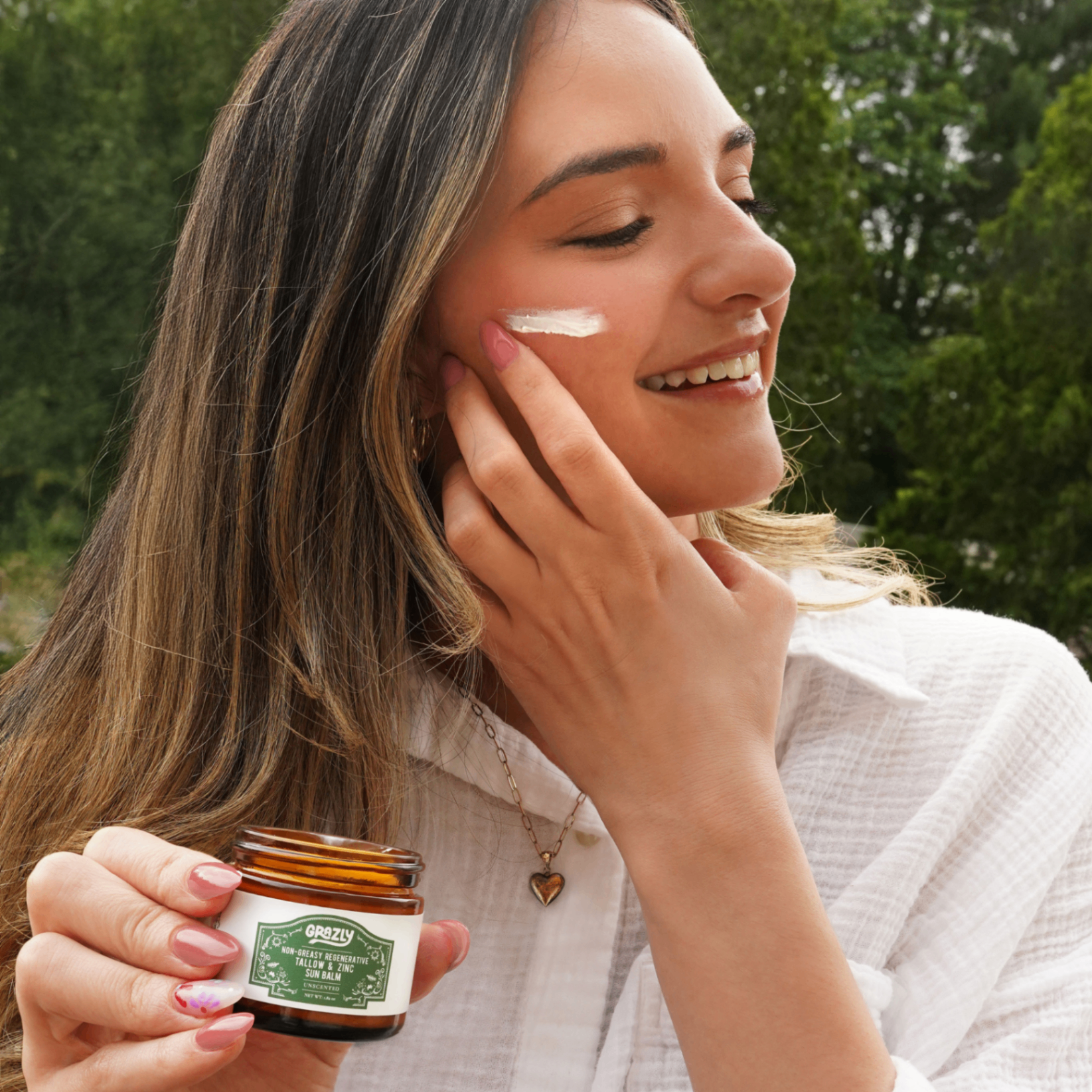Woman applying cream to her face, holding a jar, outdoors.