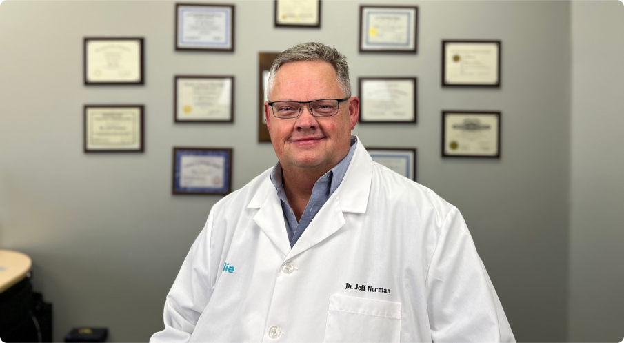 Person in a white coat with certificates on the wall behind.