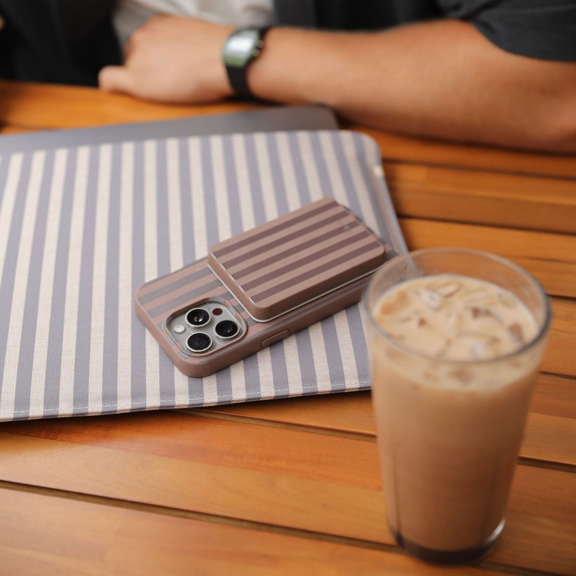 A striped phone case and mat on a wooden table next to a glass of iced coffee.