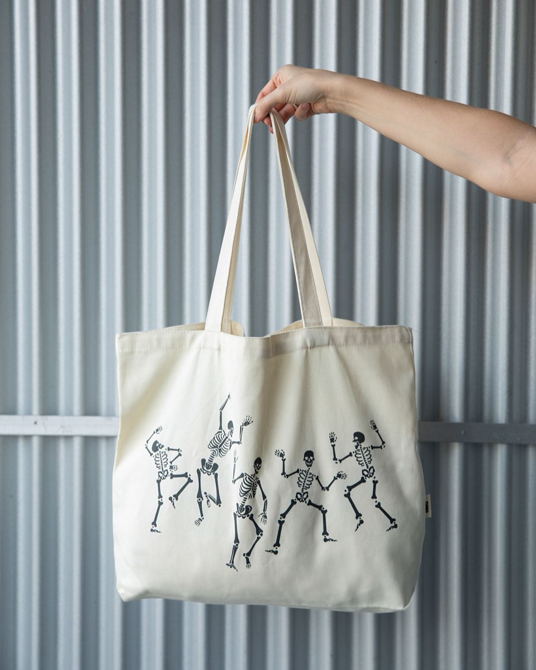 A hand holds a cream-colored tote bag featuring a black print of several dancing skeletons.