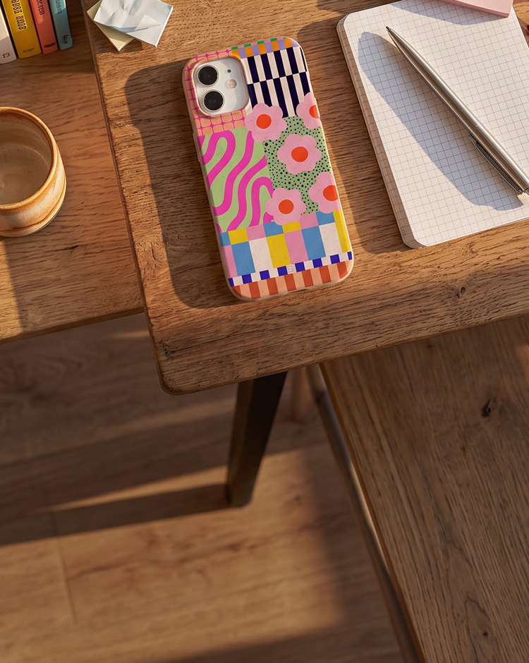A smartphone in a brightly colored, patterned case rests on a wooden desk next to a notebook and pen.