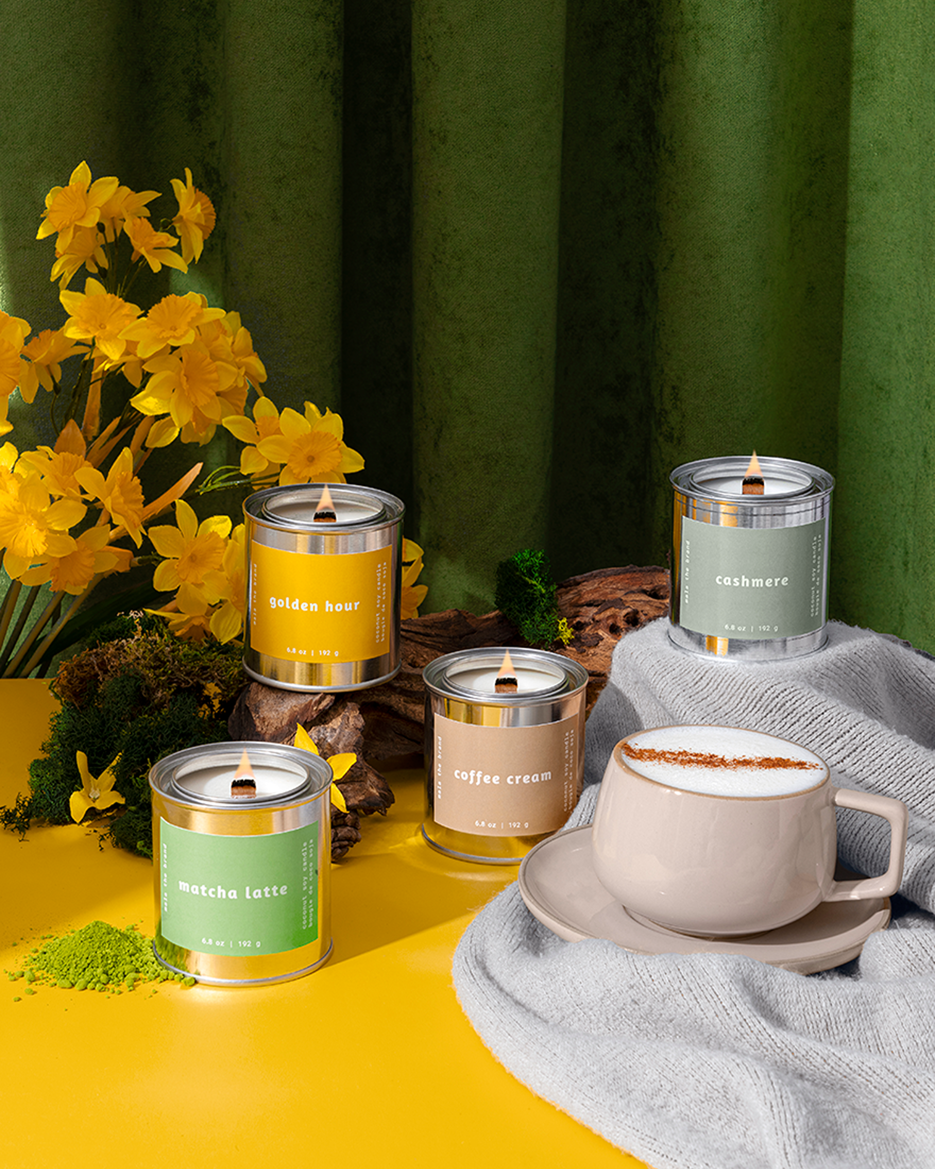 Four lit candles in tins are arranged with a cup of latte and yellow daffodils on a colorful background.