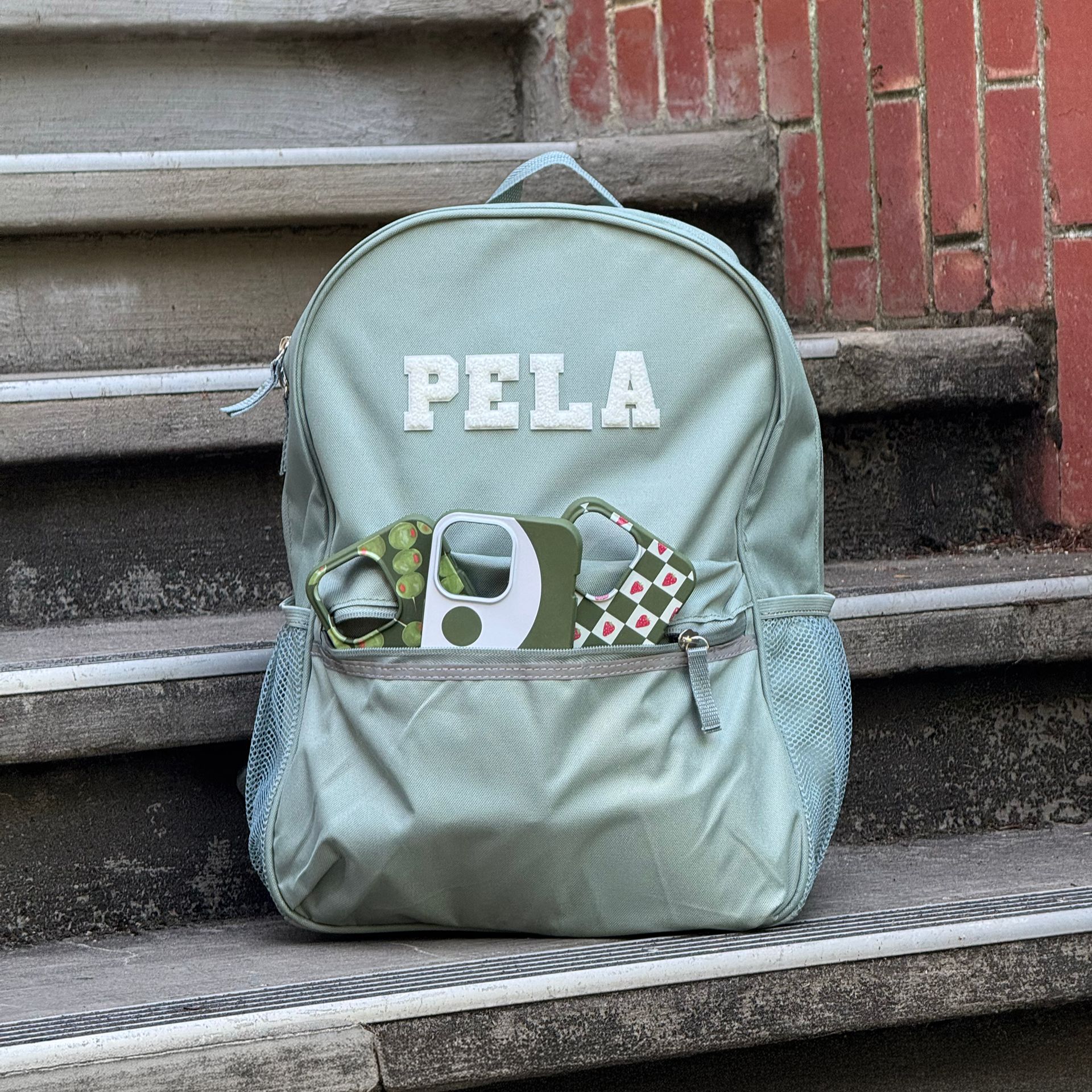 A light green PELA backpack sits on concrete steps with phone cases in its front pocket.