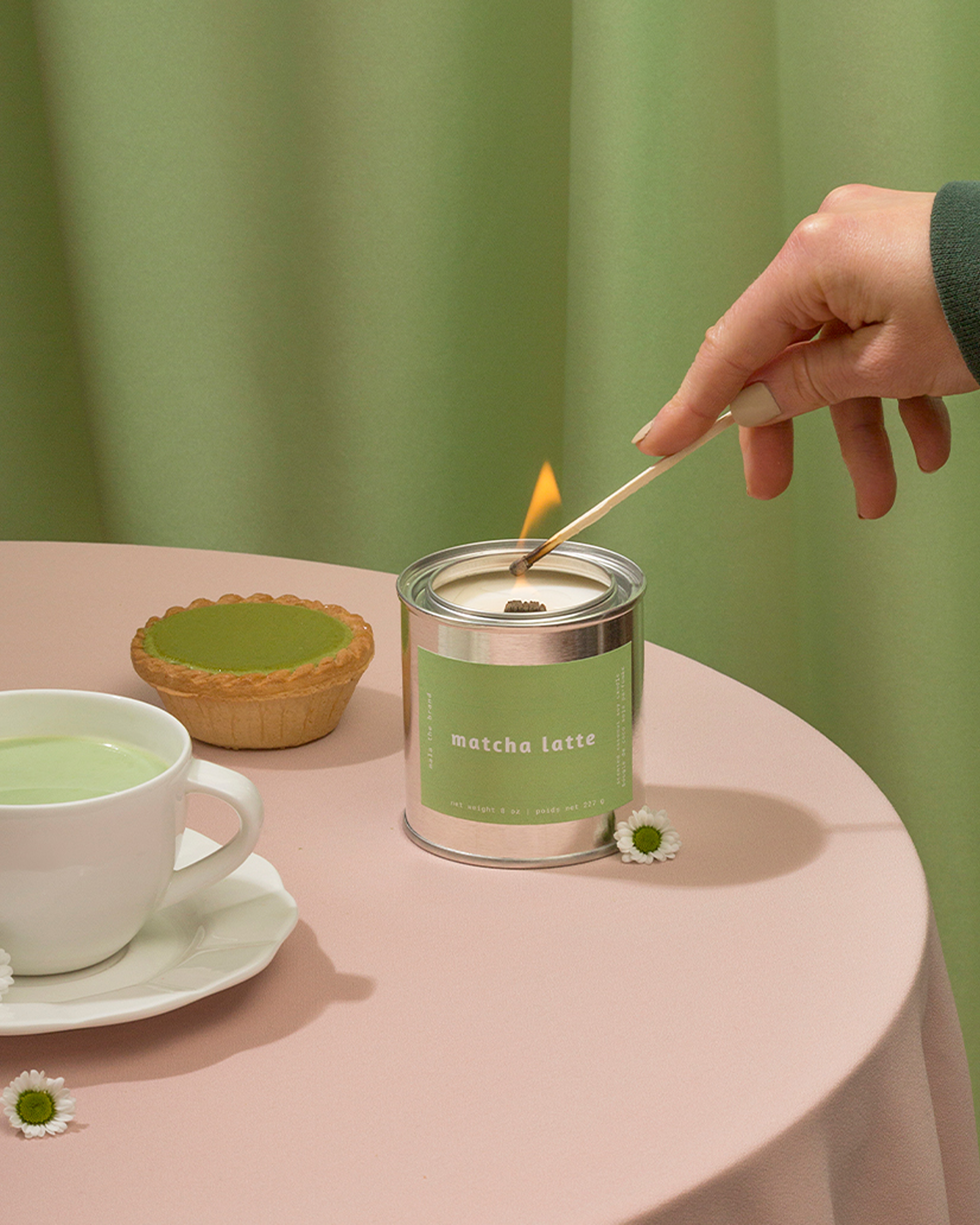 A hand lights a matcha latte candle on a pink table with a matcha drink and a tart.