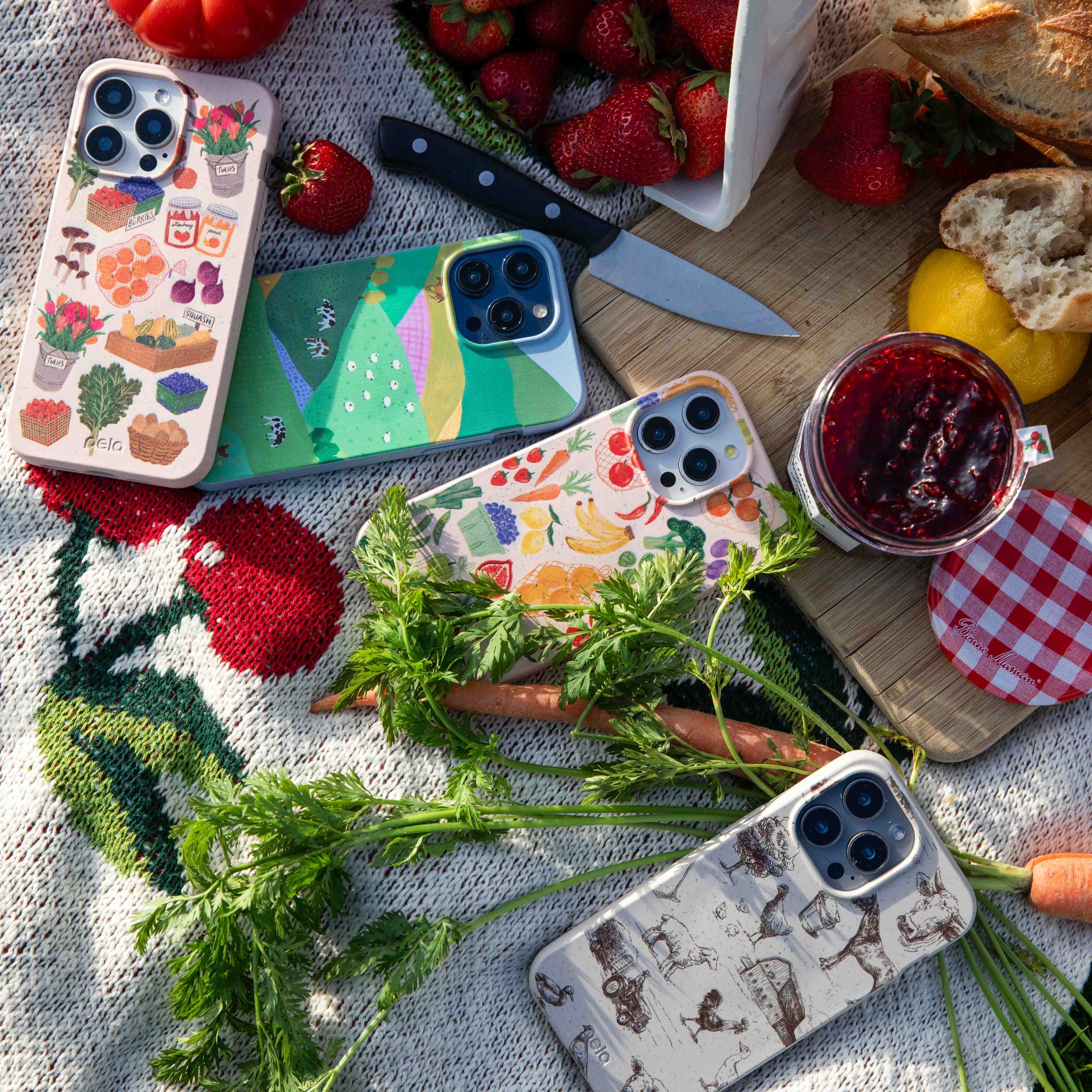 An overhead shot of four illustrated phone cases arranged on a blanket with picnic foods like strawberries and bread.
