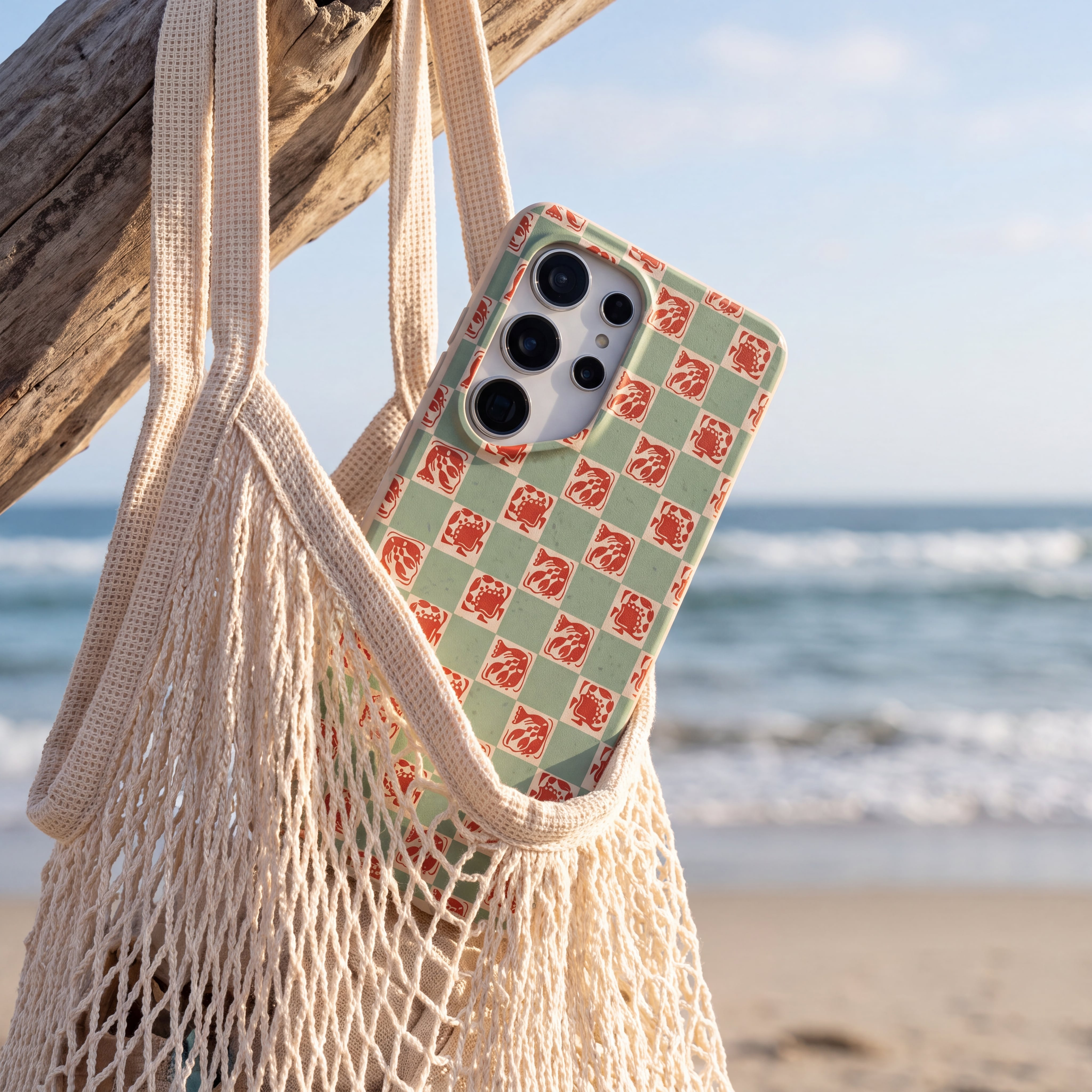 A smartphone in a green and red lobster-patterned case hangs in a net bag at the beach.