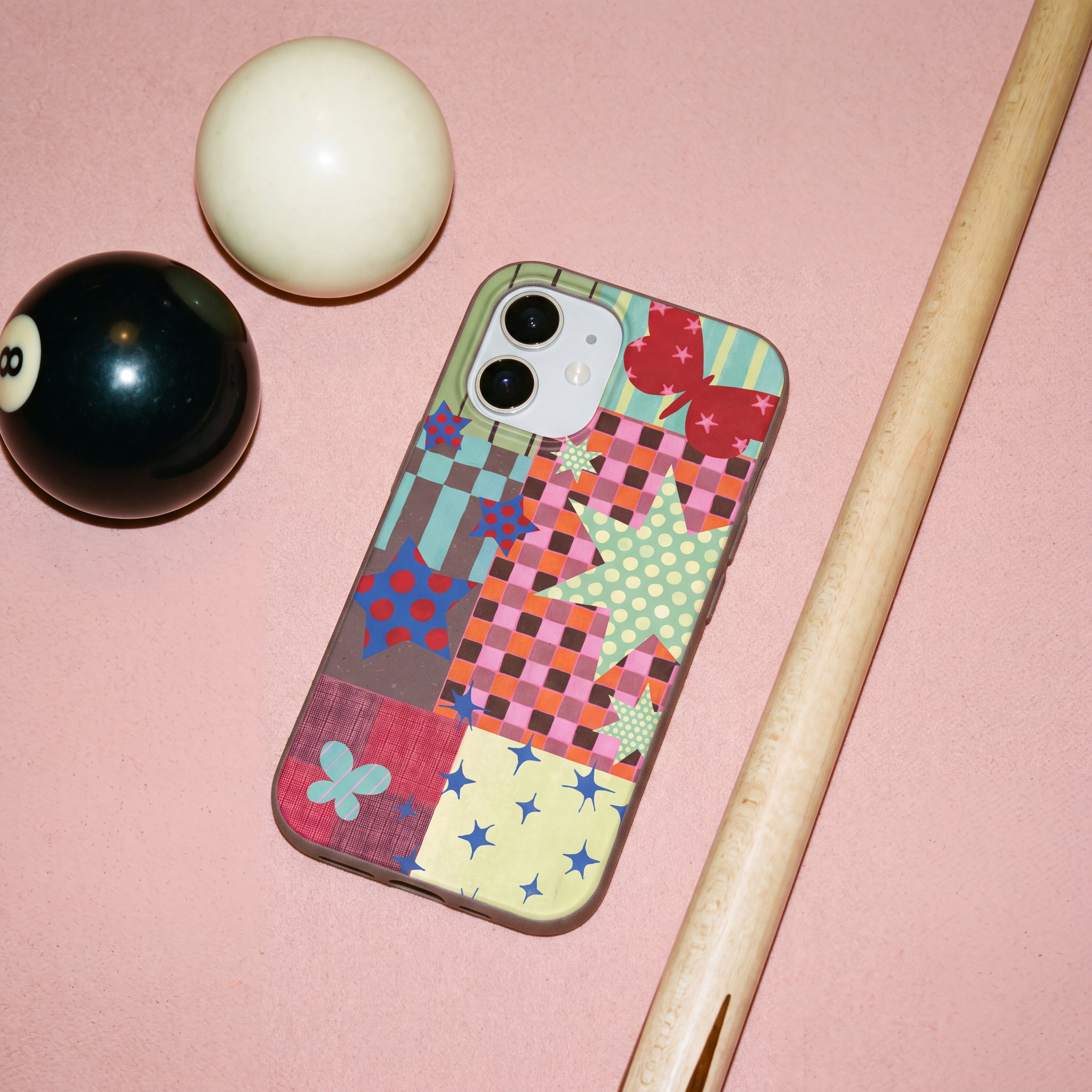 A phone in a colorful patterned case lies on a pink surface next to a pool cue and balls.