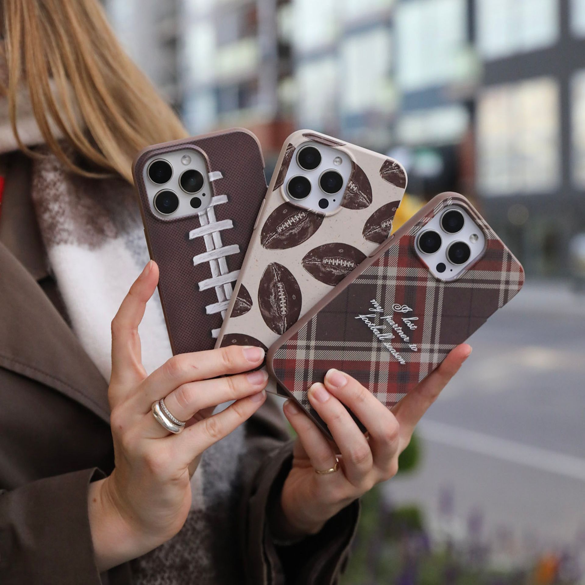 A person's hands holding up three different phone cases with football-themed designs against a blurred outdoor background.