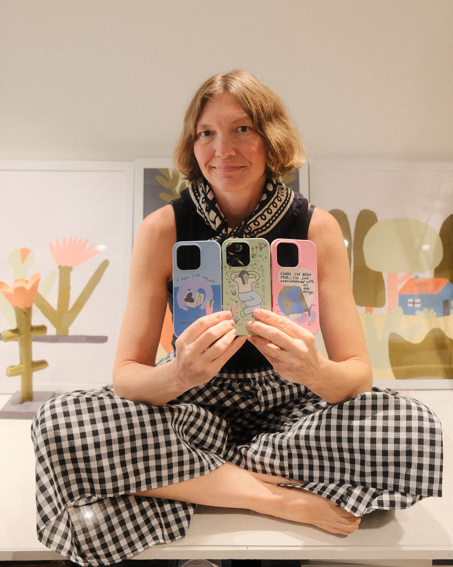 A smiling woman in a gingham skirt sits cross-legged holding up three illustrated phone cases.