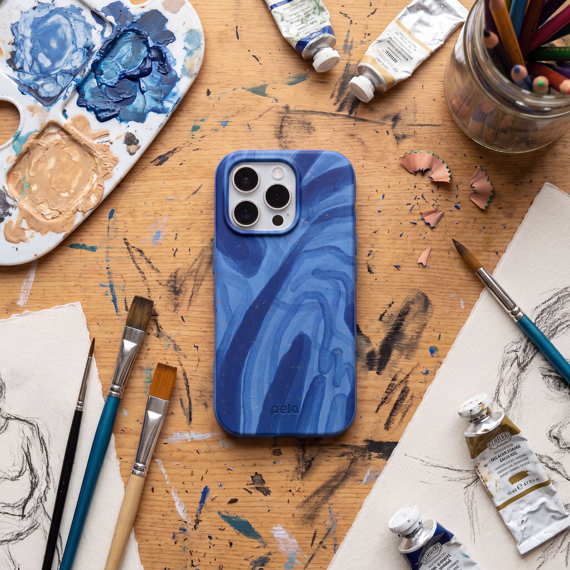 A blue patterned phone case sits on a messy artist's desk surrounded by paints, brushes, and sketches.