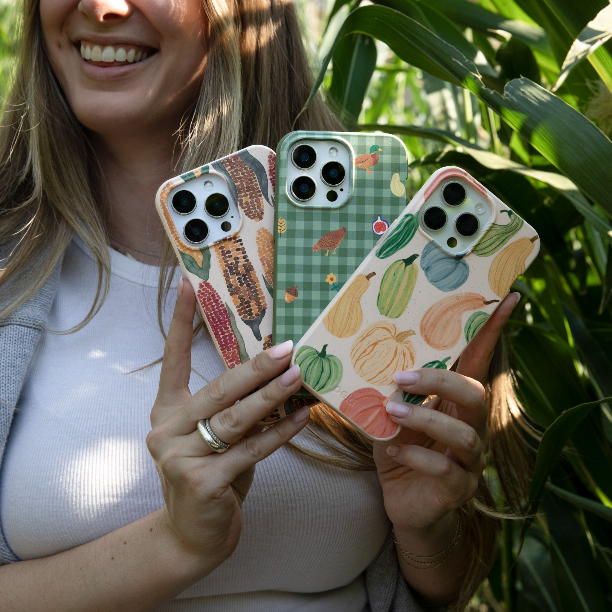 A smiling woman holds three different fall-themed phone cases while standing in a cornfield.