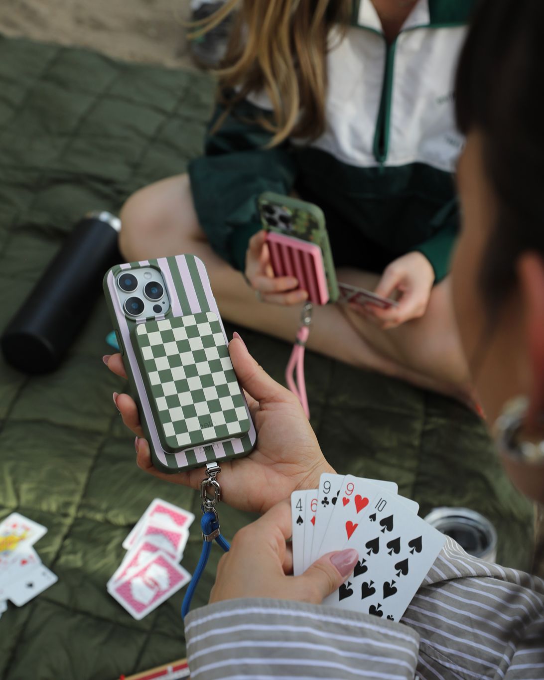Two people on a blanket playing cards, one holding a phone with a checkered green cardholder.