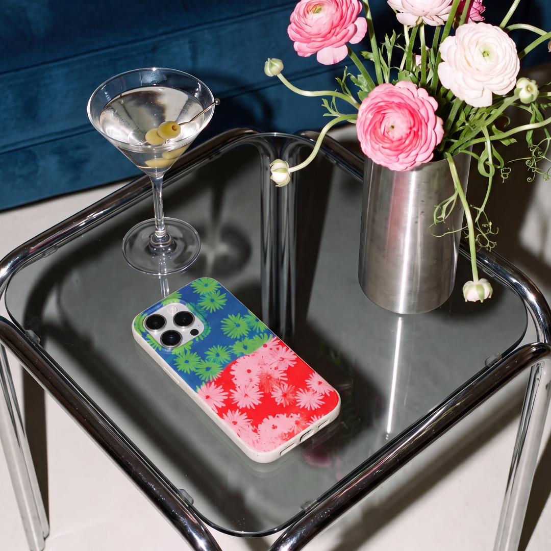 A floral phone case on a glass table next to a martini and a vase of pink ranunculus flowers.