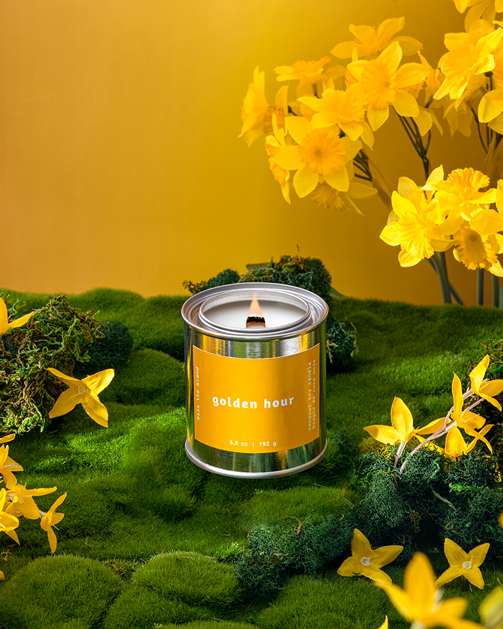 A lit 'golden hour' candle in a tin sits on green moss, surrounded by yellow daffodils.