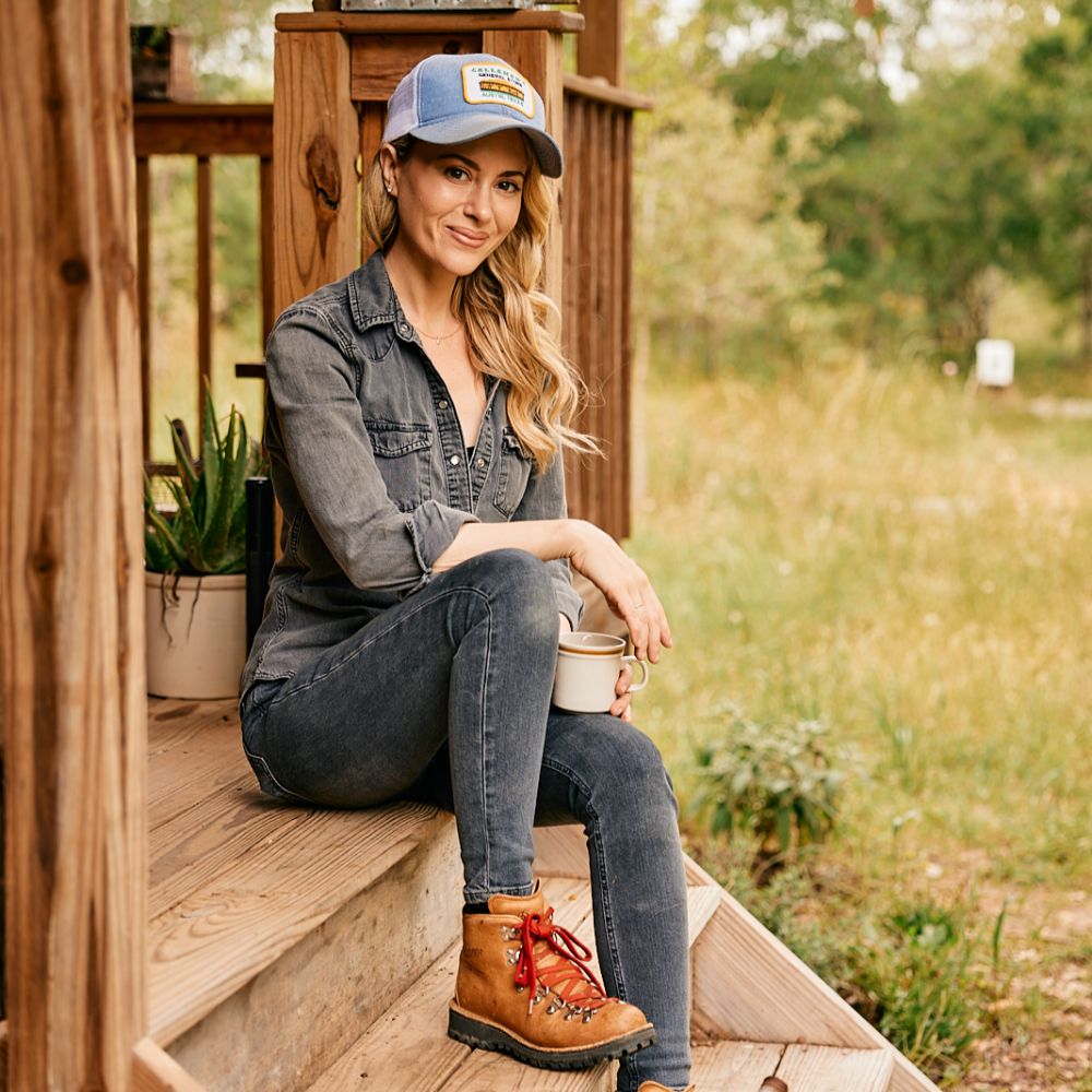 A woman in a hat and hiking boots sits on wooden steps, holding a mug and smiling.
