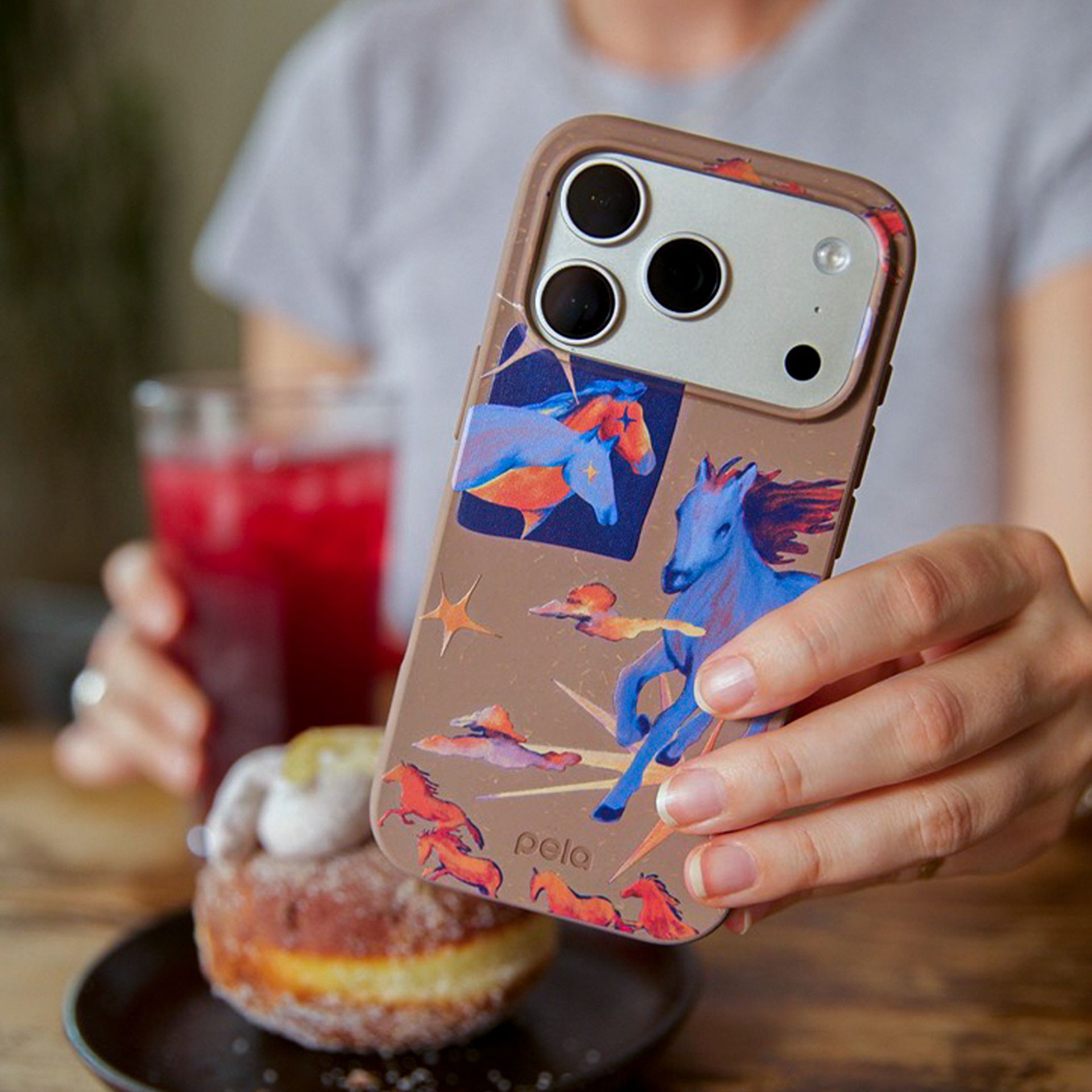 A person holds up a brown phone case with colorful horse illustrations, with food and drink behind it.