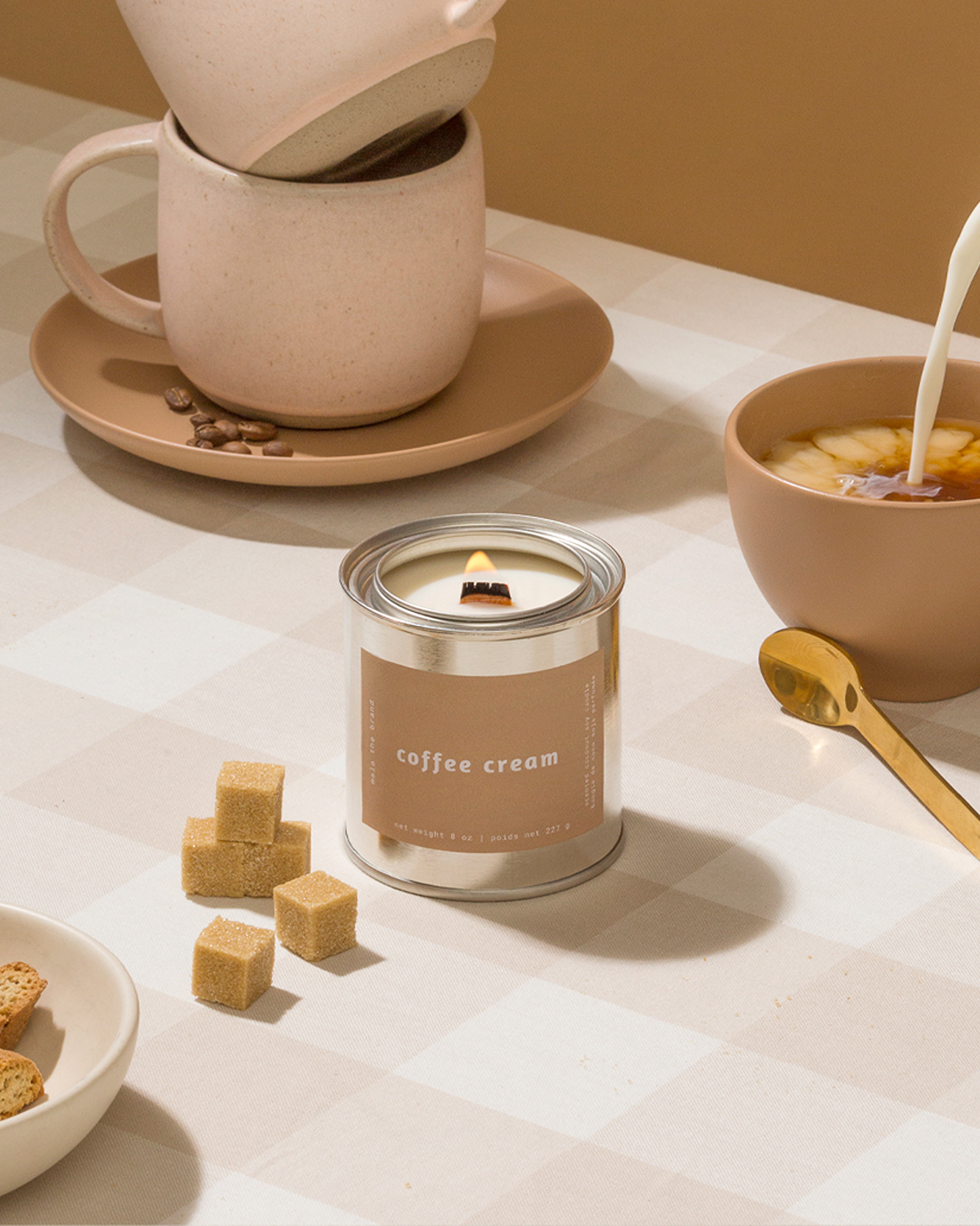 A lit 'coffee cream' candle on a checkered tablecloth with coffee mugs, sugar cubes, and coffee beans.