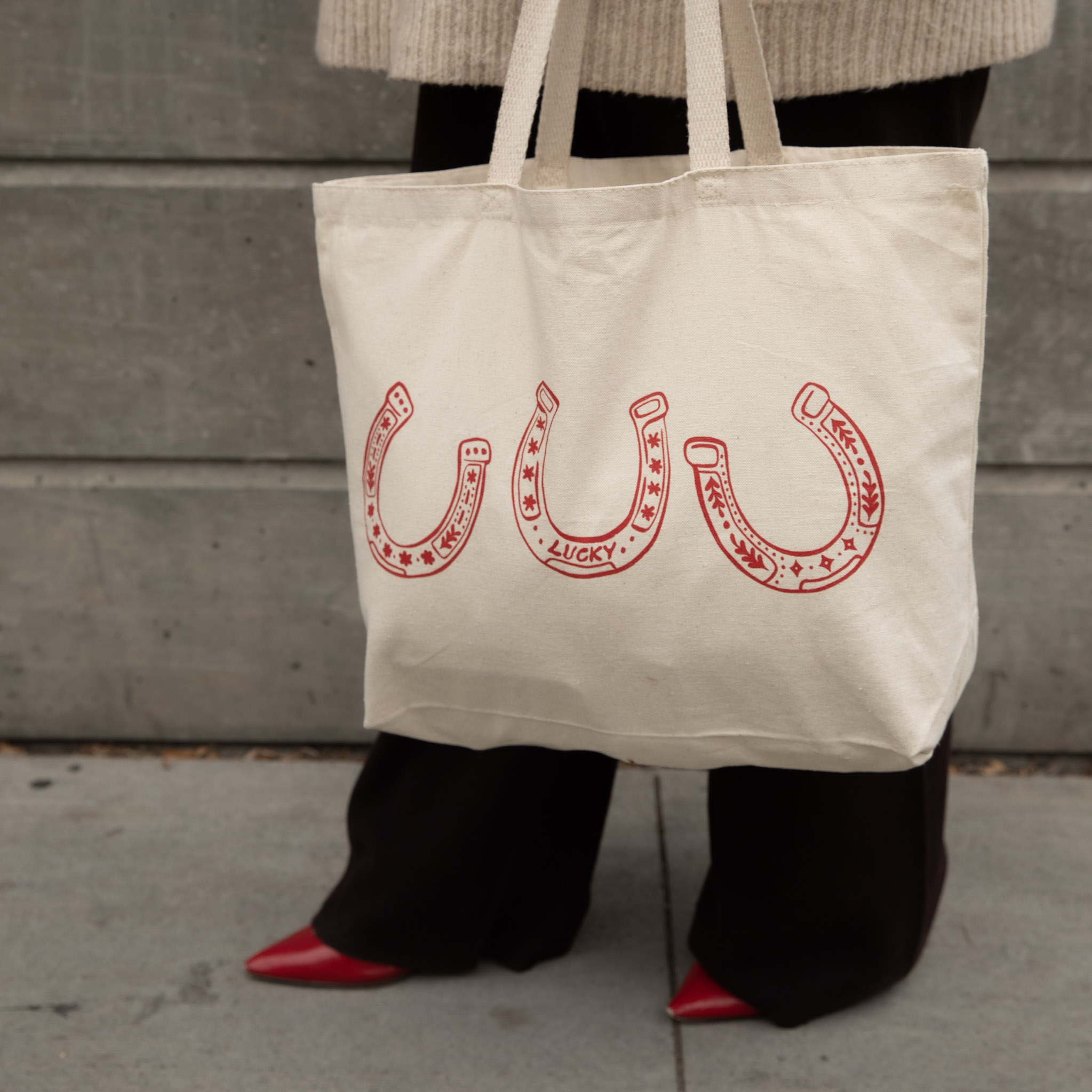 A person holds a cream tote bag with a red graphic of three lucky horseshoes.