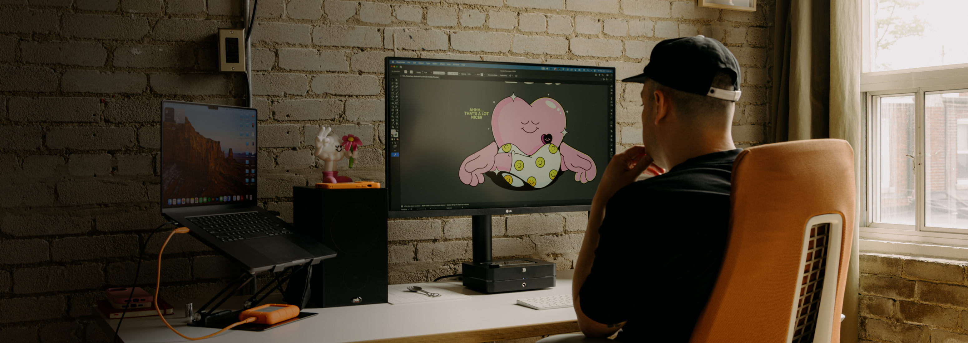 A person from behind wearing a cap works on a cartoon heart illustration on a large computer monitor.