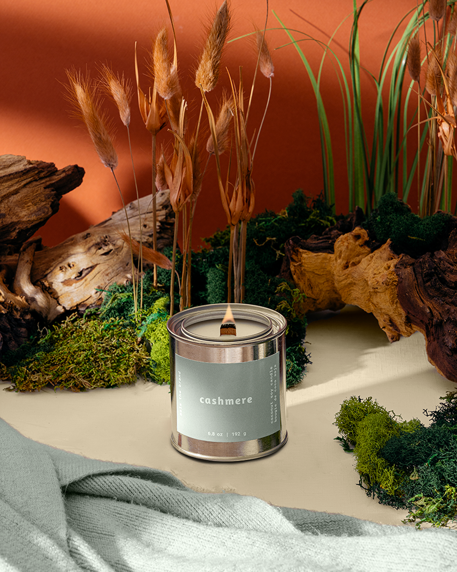 A lit 'cashmere' candle in a tin, surrounded by moss, driftwood, and dried grasses on a table.