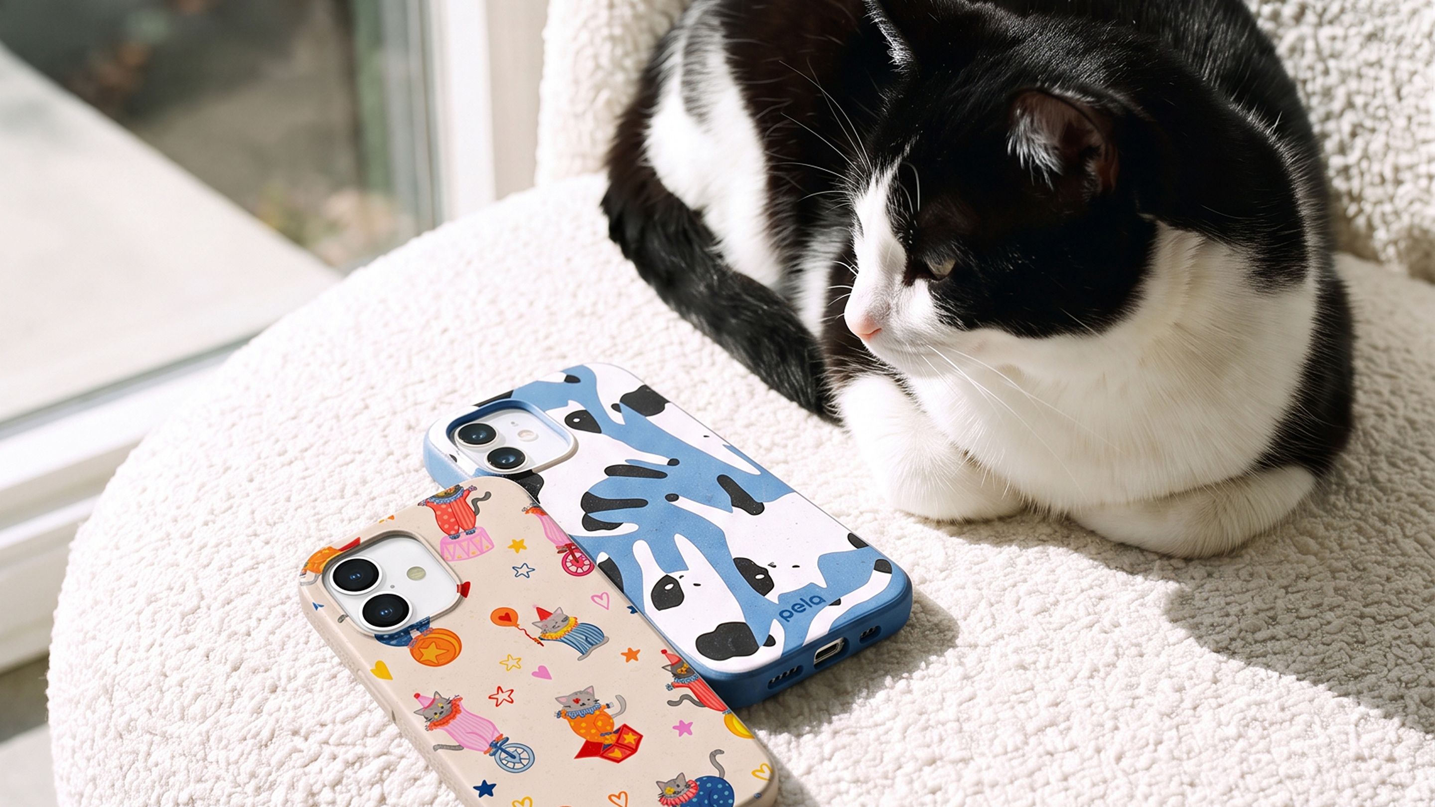 A black and white cat rests on a fluffy white surface next to two iPhones in decorative cases.
