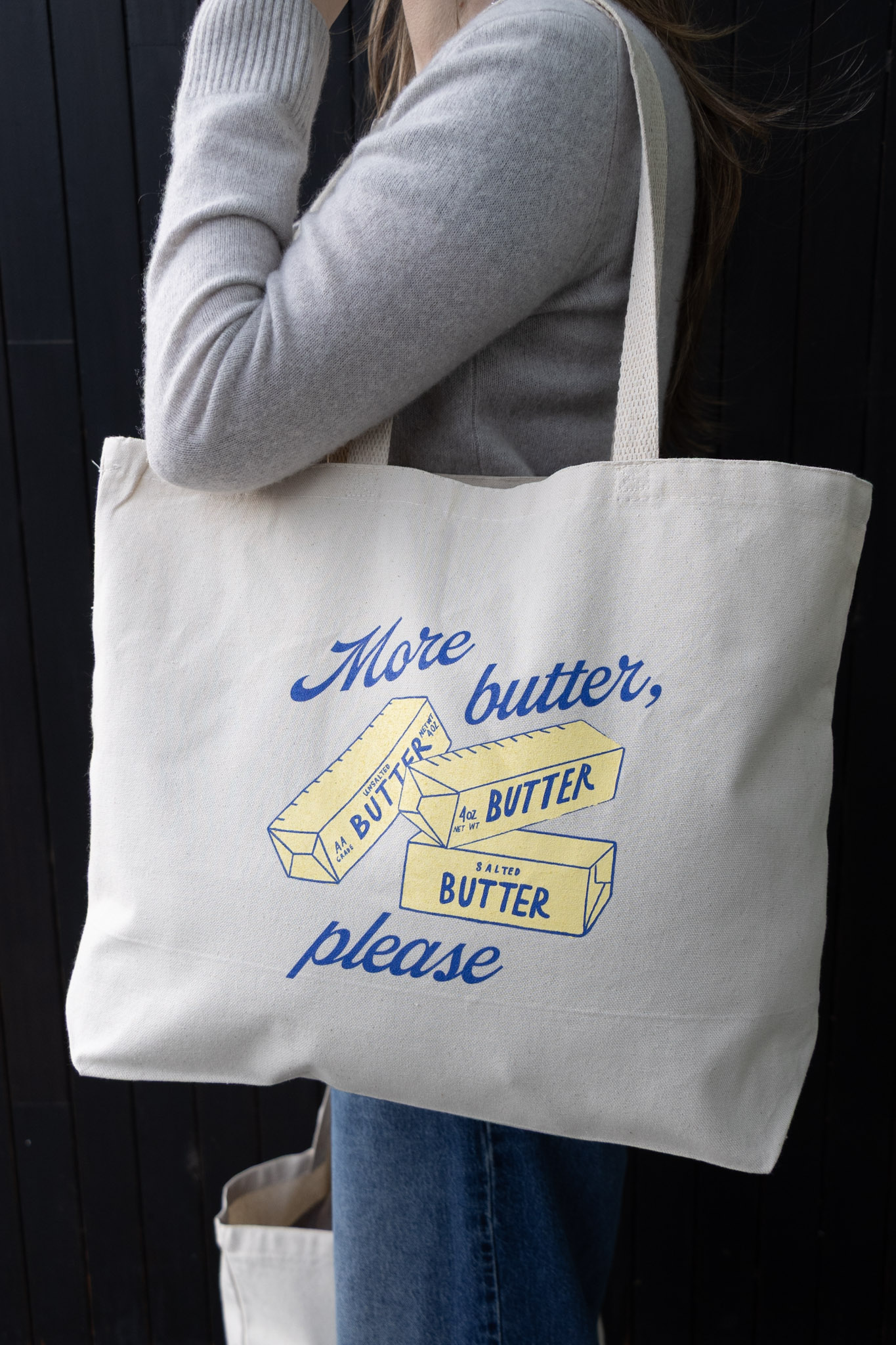 A person wearing a grey sweater holds a canvas tote bag reading 'More butter, please' with illustrations of butter.