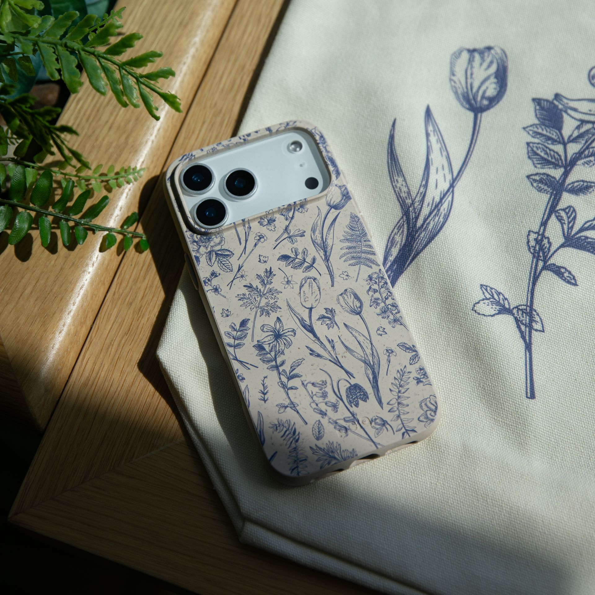 A smartphone in a case with blue floral illustrations rests on a wooden surface and a matching tote bag.