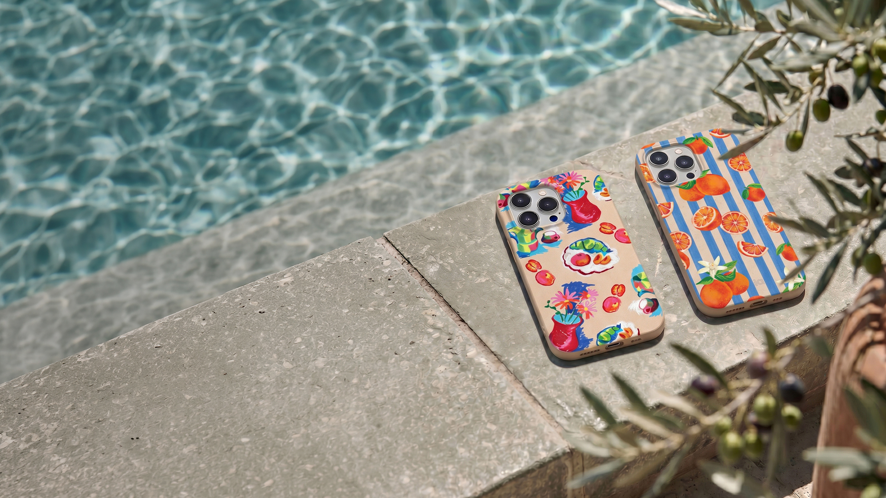 Two iPhones in colorful, patterned cases rest on a stone ledge by a shimmering swimming pool.