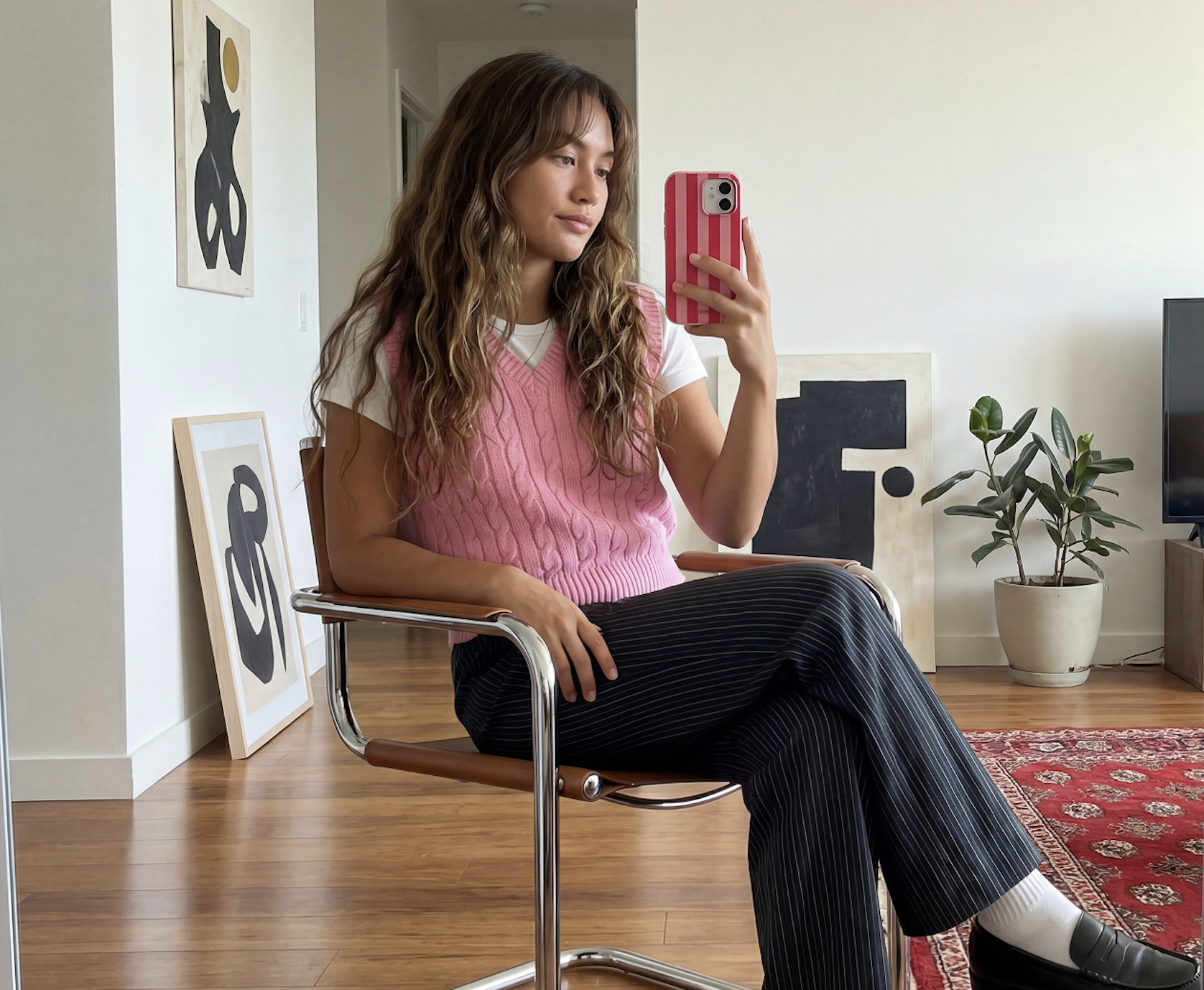 A woman in a pink sweater vest and pinstripe pants takes a mirror selfie while sitting.